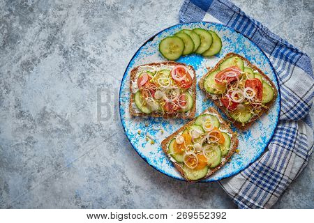 Plate with healthy toasts with cucomber, cherry tomatoes, crumbled feta cheese and radish sprouts on a black plate. Table top view. Gray stone background.