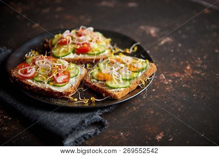 Vegetable toasts. Healthy toasts with cucomber, cherry tomatoes, crumbled feta cheese and radish sprouts on a black plate. Table top view. Dark rusty background.