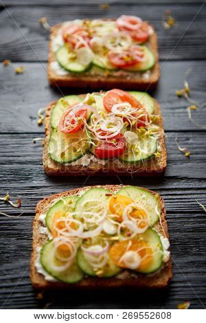 Row of three healthy toasts with onion, cucomber, cherry tomatoes, crumbled feta cheese and radish sprouts on a black plate. Table top view on black wooden background