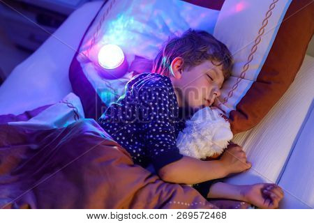 Little Preschool Kid Boy Sleeping In Bed With Colorful Lamp. School Child Dreaming And Holding Plush