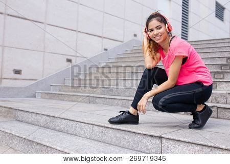 Portrait Of Smiling Young Woman Hunkering In Headphones Outdoors. Latin American Female Student List