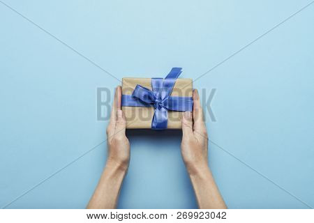 Female Hands Holding A Gift With A Blue Ribbon On A Blue Background. Concept Of A Gift For The Holid