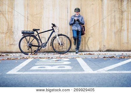 Active Senior Man With Electrobike Standing Outdoors In Town, Using Smartphone.