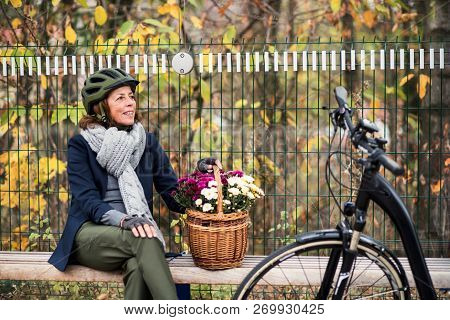 A Senior Woman With Electrobike And Flowers Sitting On A Bench Outdoors In Town.