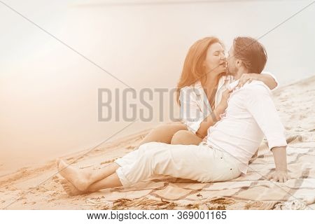 Couple In Love Gently Hugs And Kisses Sitting On Blanket In The Sand Near Water. Mature Couple In Wh