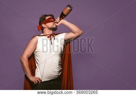 Super Antihero Man Drinks Alcohol From The Throat Of A Bottle. Isolated On Grape Purple Background W