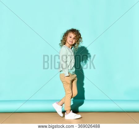 Little Curly Kid In T-shirt, Denim Jacket, Beige Pants And White Sneakers. She Smiling, Hands In Poc