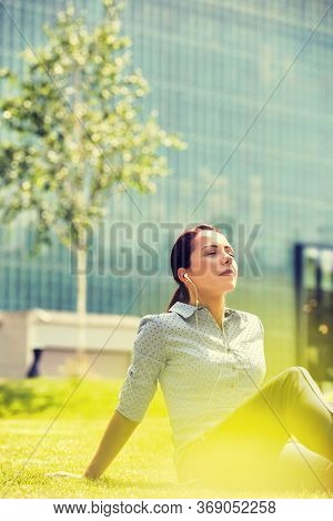 Portrait of young arrractive businesswoman sitting on the grass while listening on music with earphones on
