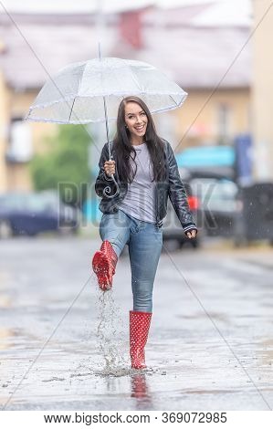 Happy Woman In Red Polka Dot Rainboots  Walks On The Street With An Umbrella On A Raidy Day.
