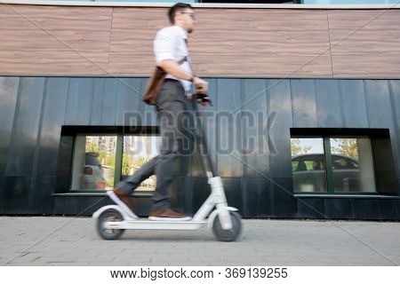Blurry young businessman on scooter moving forwards along wall of contemporary business center while going home after work