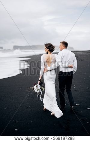 Destination Iceland Wedding. A Wedding Couple Is Walking Along The Black Beach Of Vic. Sandy Beach W