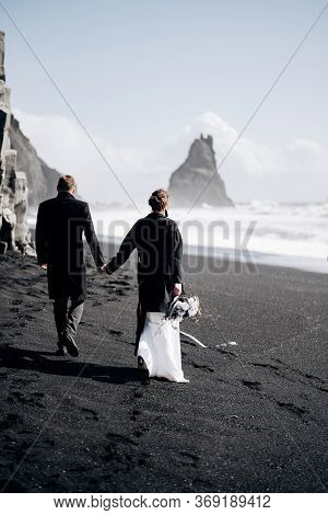 Destination Iceland Wedding. A Wedding Couple Walks Along The Sandy Black Beach Of Vik, Near The Bas