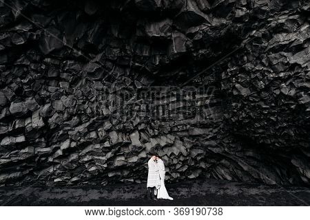 Wedding Couple Under A Rock Of Basalt Stones. The Bride And Groom On A Black Vik Beach, Sheltered By