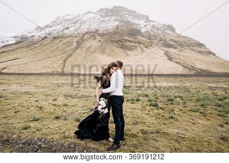 Wedding Couple On A Background Of Snowy Mountains. The Bride In A Black Dress And Groom Are Hugging 