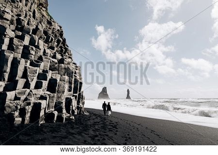 Destination Iceland Wedding. A Wedding Couple Walks Along The Sandy Black Beach Of Vik, Near The Bas