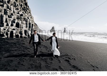 Destination Iceland Wedding. A Wedding Couple Walks Along The Sandy Black Beach Of Vik, Near The Bas