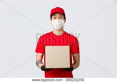 Surprised Courier In Red Uniform Cap And T-shirt, Holding Box With Client Order, Wearing Medical Mas