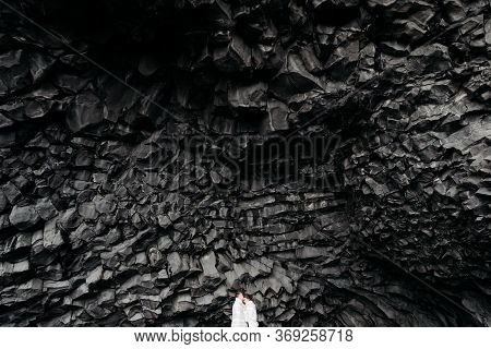 Wedding Couple Under A Rock Of Basalt Stones. The Bride And Groom On A Black Vik Beach, Sheltered By