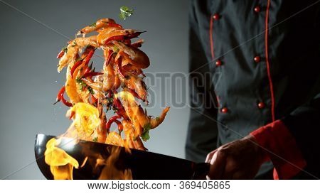 Closeup of chef throwing prawns from wok pan in fire. Fresh asian food preparation on dark background.