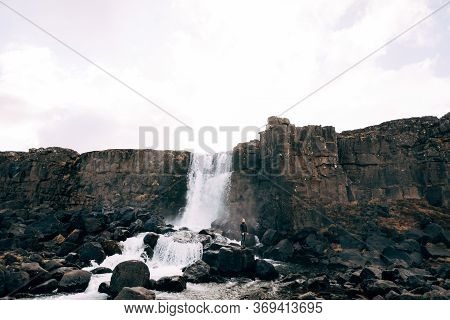 A Guy In A Black Knitted Sweater Stands On Rocks Near Ehsaraurfoss Falls, Ehsarau River, National Pa