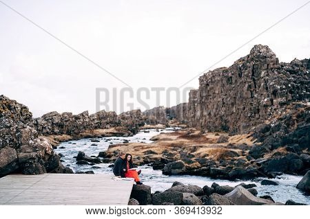 A Guy And A Girl Are Sitting Embracing On A Bridge Near Ehsaraurfoss Waterfall, Ehsarau River, Natio