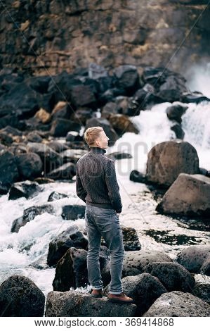 A Guy In A Black Knitted Sweater Stands On Rocks Near Ehsaraurfoss Falls, Ehsarau River, National Pa