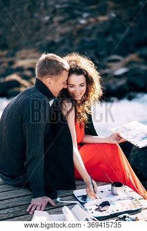A Guy Hugs A Girl Artist Who Paints A Picture In The Album With Watercolors. Near Ehsaraurfoss Water