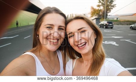 Closeup Photo Of Brunette And Blonde Smiling Girls Making Selfie On Smartphne At Car Parking