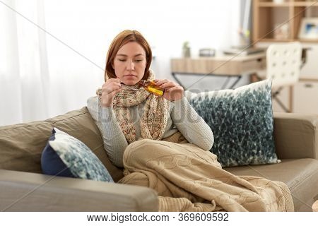 people, health and cold concept - sad sick woman in blanket and scarf pouring cough syrup or antipyretic medicine at home