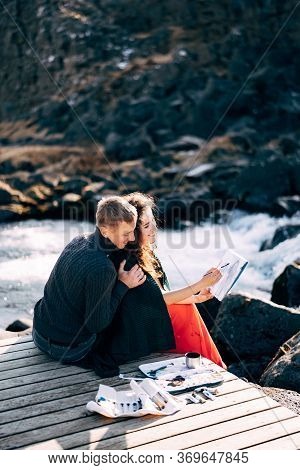 A Guy Hugs A Girl Artist Who Paints A Picture In The Album With Watercolors. Near Ehsaraurfoss Water