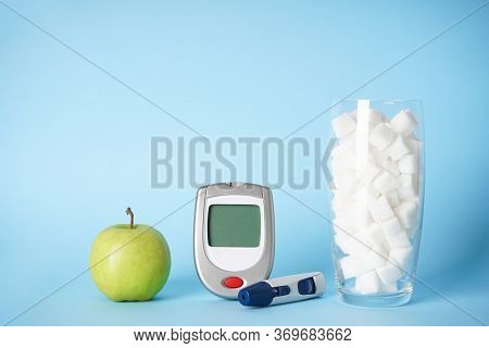 Diabetes Concept. Digital Glucometer, Lanceolate Pen, Sugar In A Glass And An Apple On A Blue Backgr