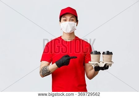 Smiling Asian Delivery Man Red Uniform Cap, Medical Mask And Gloves, Holding Client Order, Pointing 