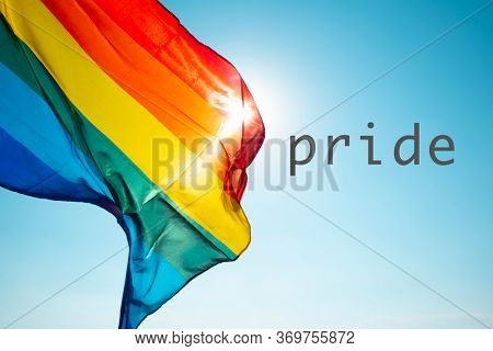 closeup of a rainbow flag waving on the blue sky, moved by the wind, with the sun in the background, and the word pride