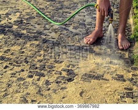 A Man Washes His Bare Feet From A Watering Hose, Standing On A Sandy Paving Stone, In The Summer Out