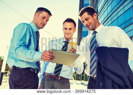 Mature attractive businessman showing and discussing plans over digital tablet with colleagues against office building