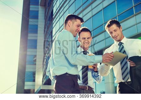 Mature attractive businessman showing and discussing plans over digital tablet with colleagues against office building