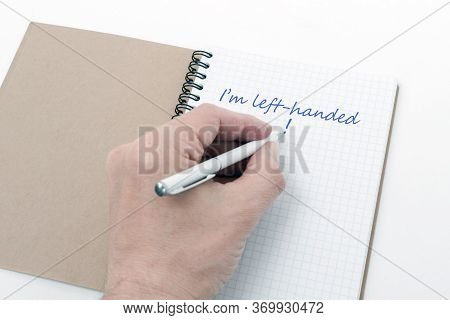 Left-handed Day Concept. Above The Open Notebook, The Left Hand With A Pen. The Inscription In The N