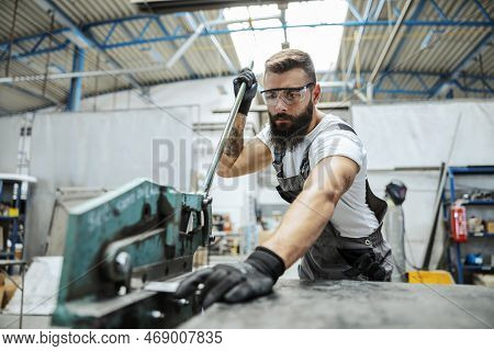 A Blue-collar Worker In A Protective Uniform Is Standing In His Workshop And Cutting Metal Parts. Wo