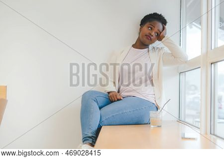 Relaxed African Woman Drinking Morning Coffee At Cafe, Sitting Near Window On Windowsill. Deamy Woma