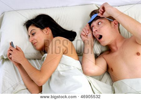 Jovem casal deitado na cama, traindo o marido usando um celular.