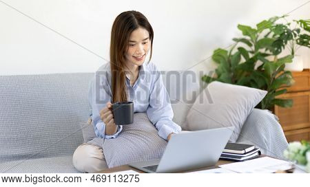 Beautiful Asian woman working on laptop and sipping coffee with smiling face in her home, Creating h