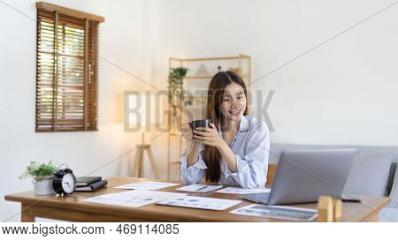 Beautiful Asian woman working on laptop and sipping coffee with smiling face in her home, Creating h
