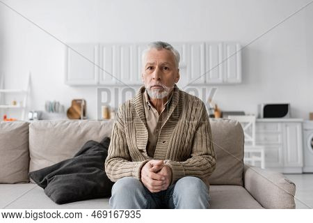Senior Grey Haired Man With Azheimers Syndrome Sitting On Couch At Home And Looking At Camera.