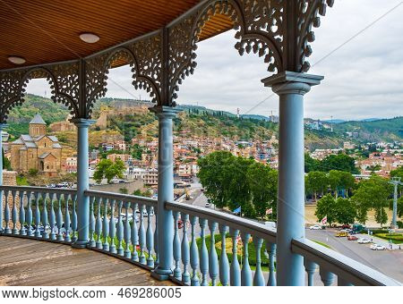 Carved Wooden Balcony With View To Metekhi Church, Europe Square And Colorful Buildings In Historica
