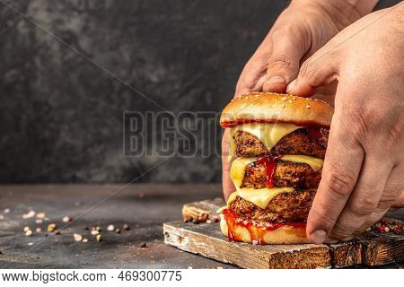 Man Holds Burger Juicy Burger With Triple Patty With Hands On Dark Background. Fast Food And Junk Fo