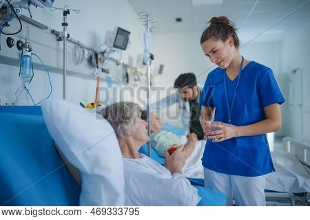 Young Nurse Giving Glass Of Water To Senior Patient At Hospital Ward.