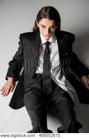 High Angle View Of Fashionable Model In Oversize Shirt And Black Pantsuit Sitting On Grey Background