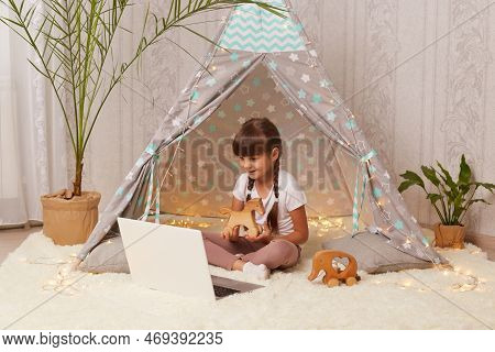 Indoor Shot Of Satisfied Delighted Little Girl With Pigtails Lying In Peetee Tent With Laptop And Ha