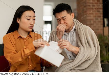 Loving Asian Woman Taking Care Of Sick Husband, Wife Giving Nupkins To Sick Man, Sitting On Sofa At 