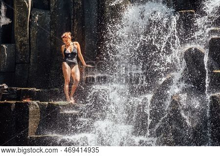Caucasian Woman In A Black Swimsuit Refreshing Under Falling Water Streams Flowing On Black Volcanic